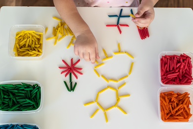 Hands of a child making a picture of flowers and the sun with noodles.